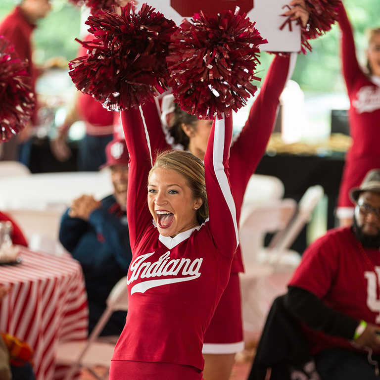A blonde woman wearing a red Indiana University cheerleader uniform raises a red pom pom 